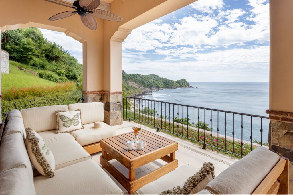 http://Un%20acogedor%20patio%20con%20asientos%20acolchados%20y%20una%20mesa%20de%20madera%20ofrece%20una%20vista%20panorámica%20del%20océano%20con%20acantilados%20rocosos,%20exuberante%20vegetación%20y%20un%20cielo%20parcialmente%20nublado.%20Un%20ventilador%20de%20techo%20cuelga%20sobre%20él,%20y%20el%20espacio%20está%20enmarcado%20por%20columnas%20y%20una%20barandilla%20de%20hierro%20forjado.