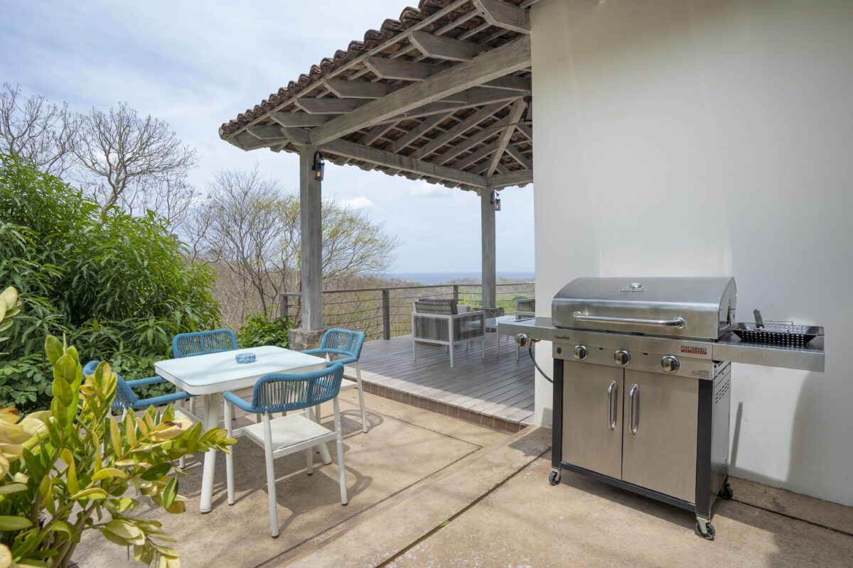 http://Patio%20al%20aire%20libre%20con%20parrilla%20a%20gas,%20una%20mesa%20blanca%20con%20cuatro%20sillas%20azules%20y%20una%20terraza%20de%20madera%20cubierta%20con%20vista%20a%20árboles%20y%20colinas%20distantes%20bajo%20un%20cielo%20nublado.