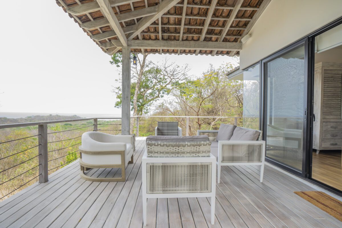 http://Un%20moderno%20patio%20al%20aire%20libre%20con%20sillas%20blancas,%20un%20sofá%20y%20una%20pequeña%20mesa%20en%20una%20terraza%20de%20madera,%20cubierta%20por%20un%20techo%20de%20tejas,%20con%20vista%20a%20los%20árboles%20y%20las%20colinas%20distantes,%20junto%20a%20una%20puerta%20corrediza%20de%20vidrio.