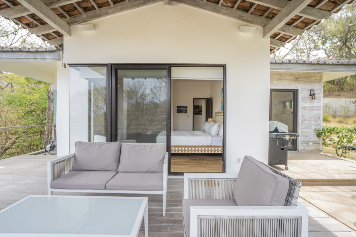 http://Un%20moderno%20patio%20exterior%20con%20dos%20sillas%20grises%20con%20cojines%20y%20una%20mesa%20de%20centro%20de%20cristal.%20Unas%20puertas%20corredizas%20de%20cristal%20conducen%20a%20un%20dormitorio%20con%20una%20cama%20tendida.%20Hay%20una%20parrilla%20y%20plantas%20visibles%20en%20un%20patio%20separado%20a%20la%20derecha.