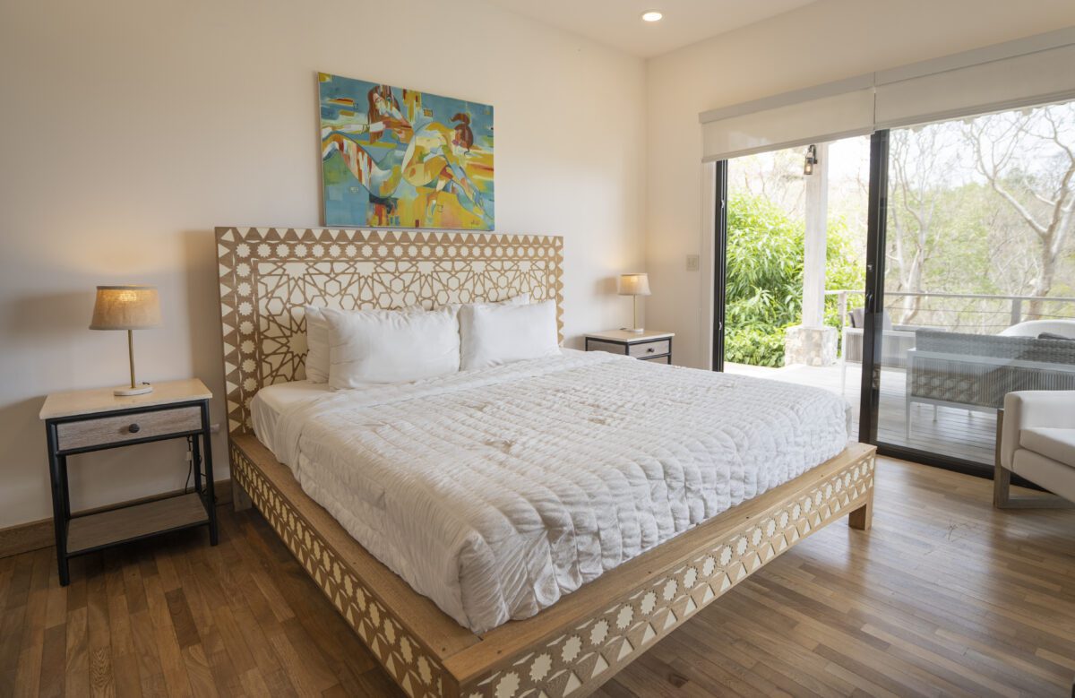 http://Un%20dormitorio%20luminoso%20con%20una%20cama%20grande%20con%20marco%20de%20madera%20decorativo,%20ropa%20de%20cama%20blanca,%20dos%20mesitas%20de%20noche%20con%20lámparas,%20un%20cuadro%20colorido%20sobre%20la%20cama%20y%20puertas%20de%20vidrio%20que%20conducen%20a%20un%20balcón%20con%20vegetación%20exterior.