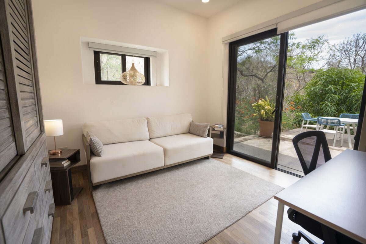http://Una%20luminosa%20oficina%20en%20casa%20con%20un%20sofá%20blanco,%20un%20escritorio%20de%20madera%20con%20silla%20y%20una%20gran%20puerta%20corrediza%20de%20vidrio%20que%20da%20a%20un%20patio%20con%20asientos%20al%20aire%20libre%20y%20exuberante%20vegetación.%20Una%20pequeña%20lámpara%20y%20una%20ventana%20aportan%20luz%20adicional%20a%20la%20habitación.