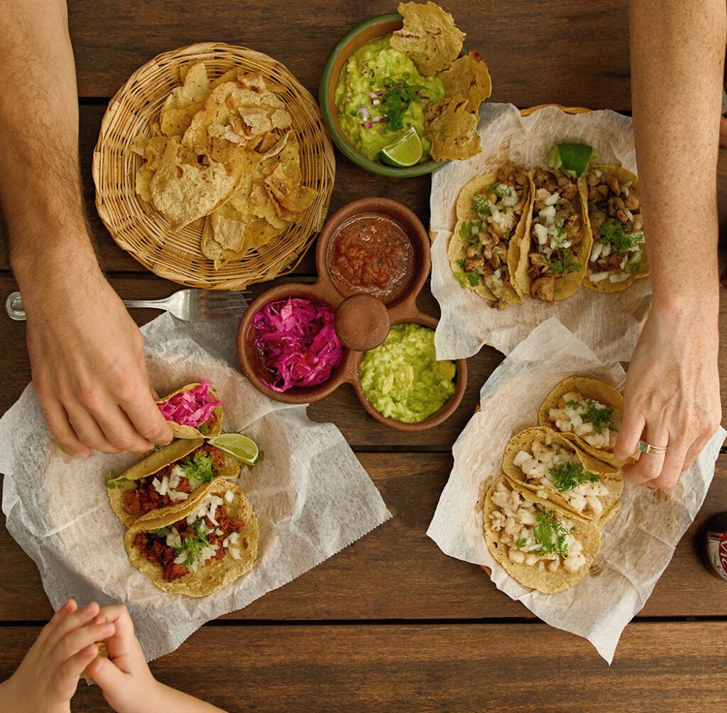 http://Tres%20personas%20comparten%20tacos,%20totopos,%20guacamole,%20salsa%20y%20cebollas%20encurtidas%20en%20una%20mesa%20de%20madera.%20Dos%20adultos%20extienden%20la%20mano%20hacia%20la%20comida%20mientras%20un%20niño%20descansa%20cerca.%20La%20comida%20se%20sirve%20en%20papel%20pergamino.