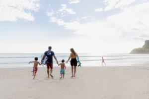 Una familia de cuatro, vista de espaldas, camina hacia el océano por una playa de arena, cargando tablas de surf. Otro niño corre delante cerca del agua. El cielo está parcialmente nublado.