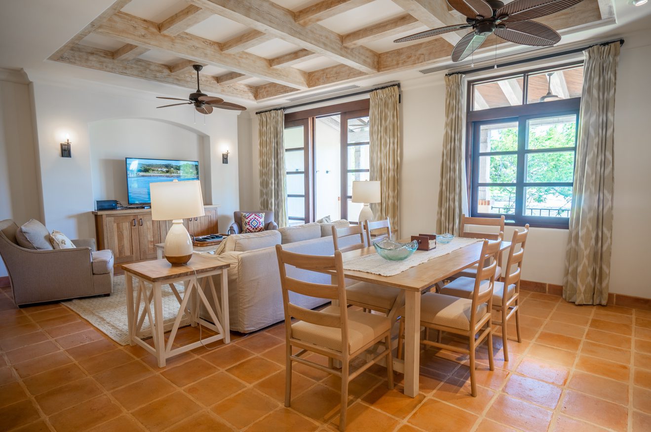 http://Sala%20de%20estar%20y%20comedor%20abiertos%20con%20vigas%20de%20madera%20en%20el%20techo,%20piso%20de%20baldosas%20de%20terracota,%20sofás%20beige,%20una%20mesa%20de%20comedor%20de%20madera%20con%20seis%20sillas,%20grandes%20ventanales%20con%20cortinas%20y%20TV%20en%20una%20consola%20en%20la%20esquina.