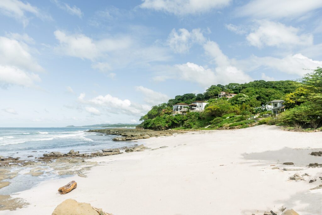 A sandy beach with scattered rocks and gentle waves, bordered by lush green hills with modern houses nestled among the trees under a partly cloudy sky.