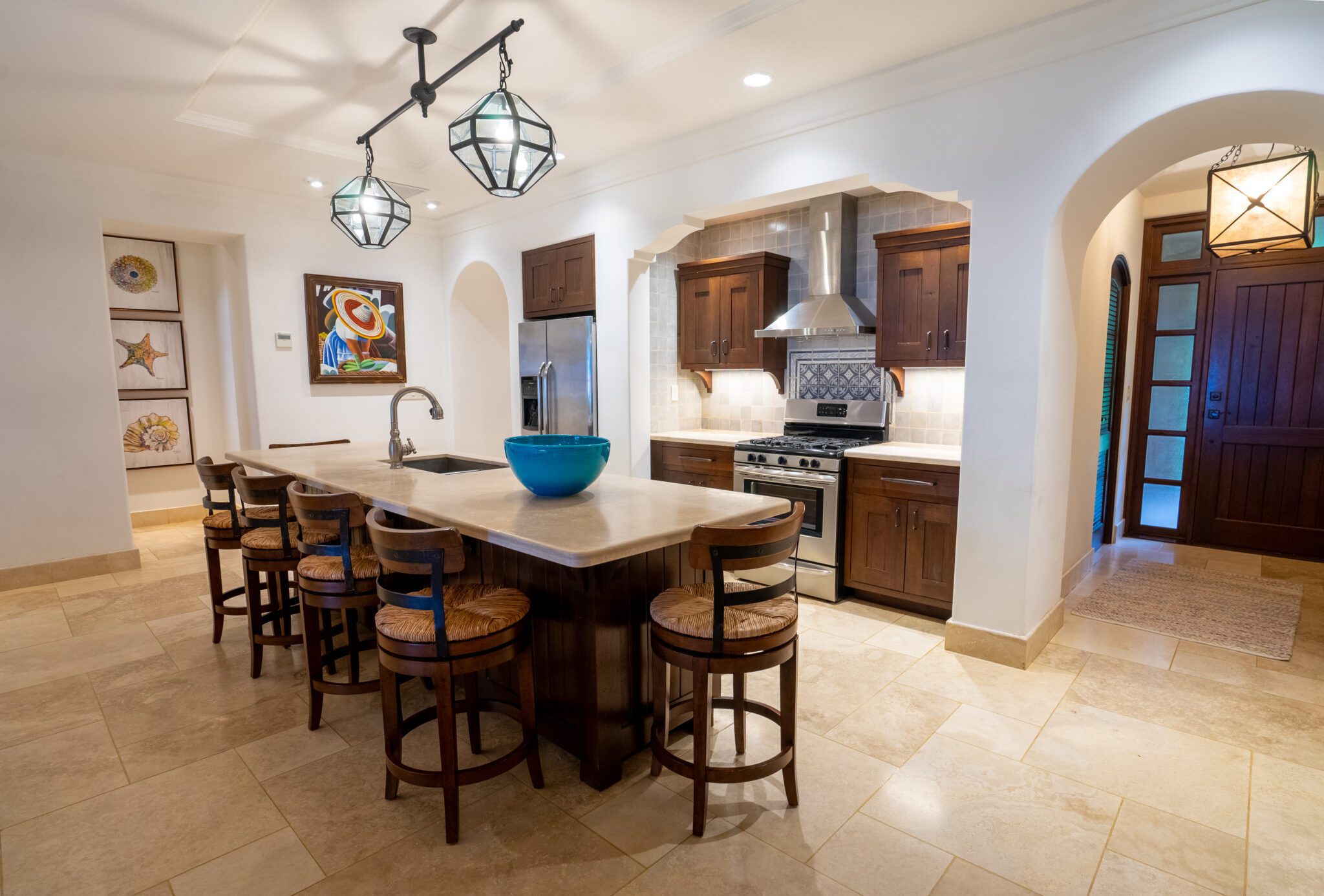 http://Una%20cocina%20espaciosa%20y%20moderna%20con%20una%20gran%20isla,%20cinco%20taburetes%20de%20madera,%20electrodomésticos%20de%20acero%20inoxidable,%20gabinetes%20de%20madera%20oscura%20y%20lámparas%20colgantes%20decorativas.%20Un%20cuenco%20azul%20se%20encuentra%20en%20la%20isla%20y%20hay%20obras%20de%20arte%20en%20las%20paredes.