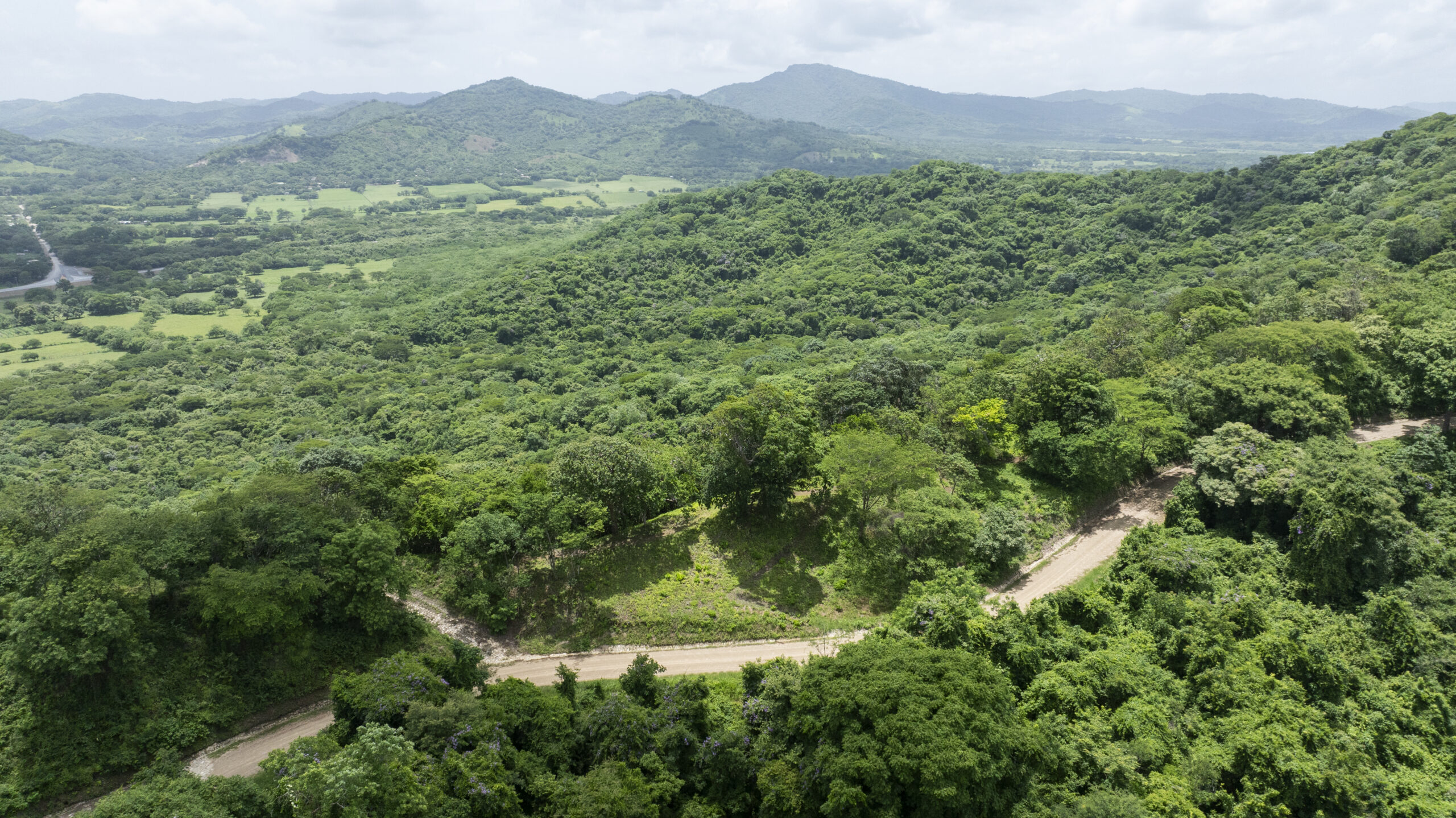 http://A%20winding%20dirt%20road%20curves%20through%20dense,%20lush%20green%20forested%20hills,%20with%20distant%20mountains%20and%20open%20fields%20visible%20under%20a%20cloudy%20sky.