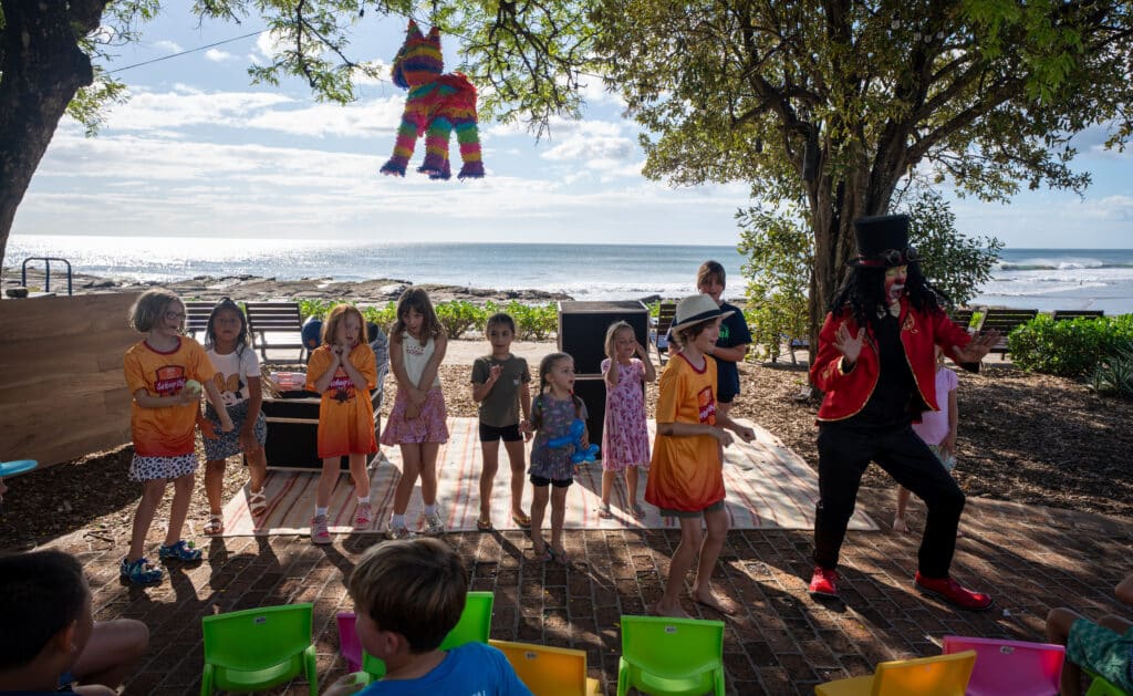A group of children stand in a row outdoors near the beach, celebrating Easter Magic with a performer. A colorful piñata hangs above them, and empty vibrant chairs are in the foreground, capturing the festive spirit of Easter Week.