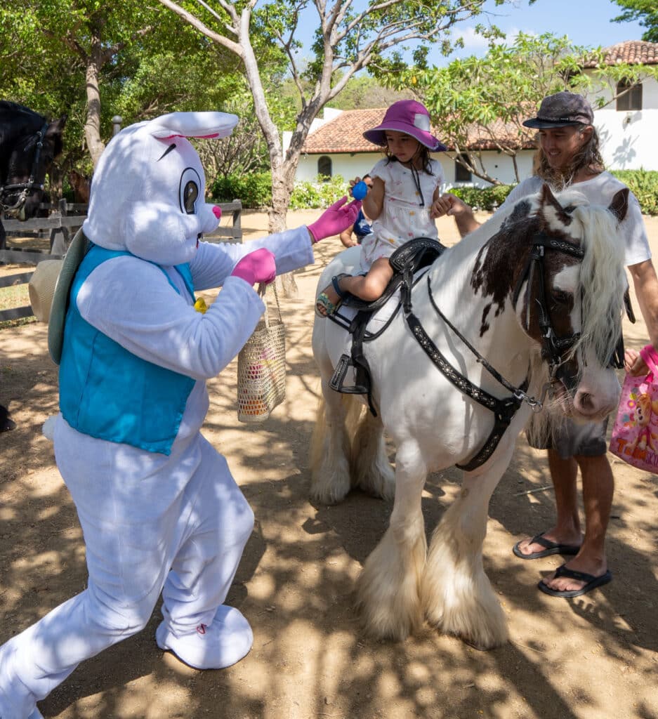 A person in a white bunny costume hands a blue egg to a young girl wearing a hat, who is sitting on a horse. An adult holds the reins nearby as Easter Magic fills the air, with trees and buildings visible in the background.