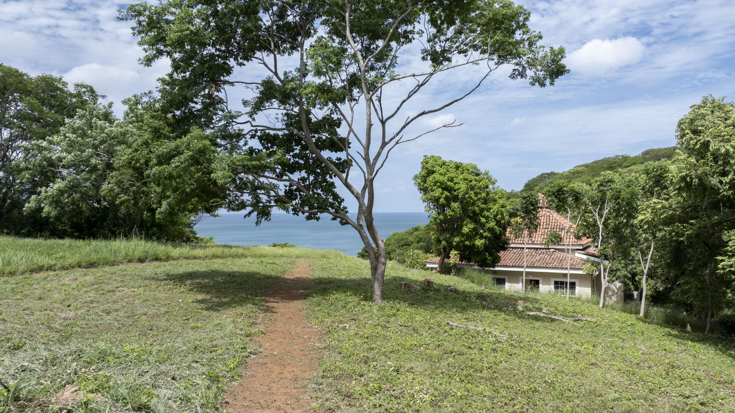 http://A%20dirt%20path%20leads%20through%20a%20grassy%20area%20with%20trees,%20toward%20a%20view%20of%20the%20ocean.%20A%20house%20with%20a%20red-tiled%20roof%20is%20partly%20visible%20on%20the%20right,%20surrounded%20by%20lush%20greenery%20under%20a%20partly%20cloudy%20sky.