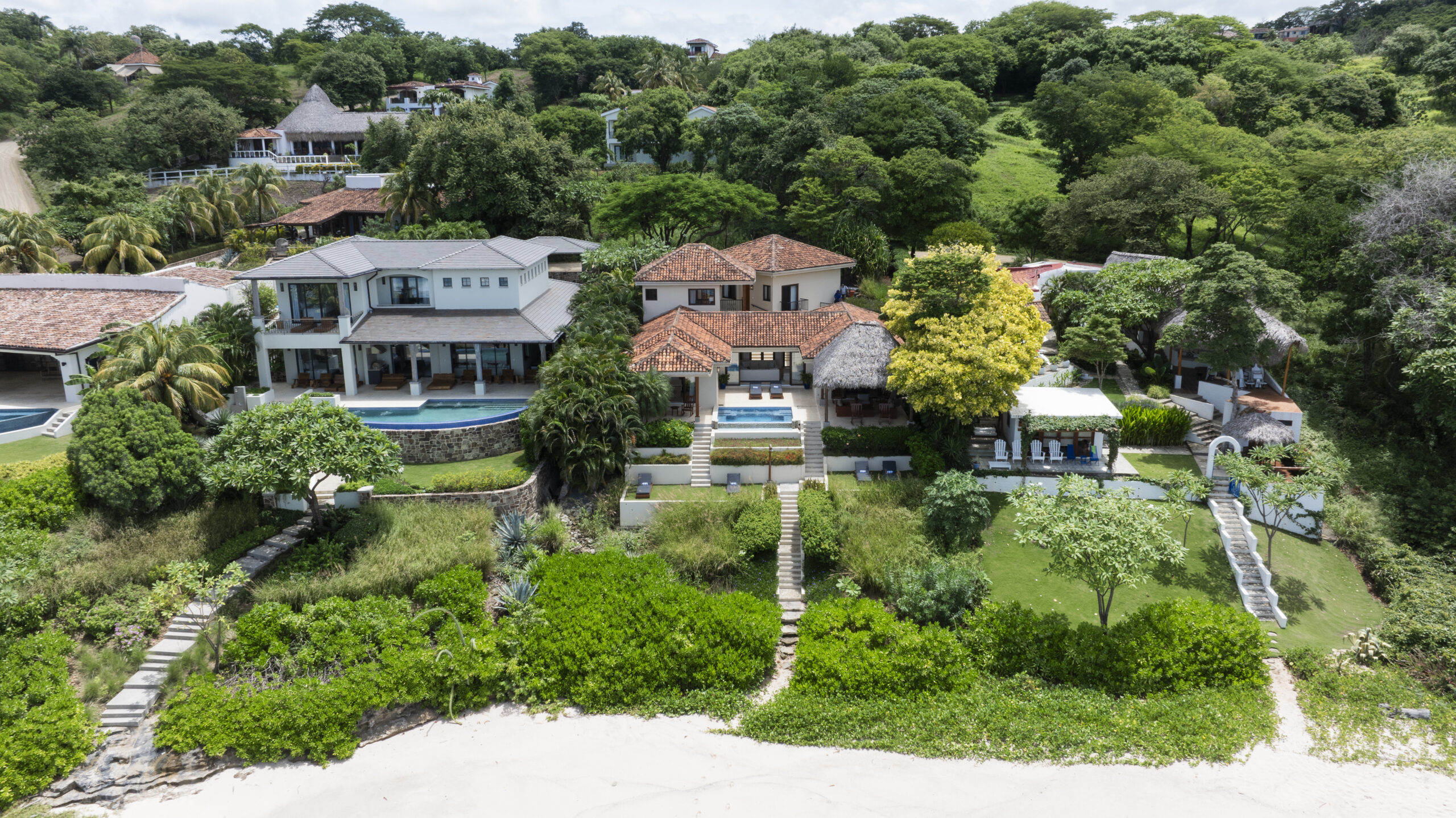 http://Aerial%20view%20of%20two%20luxury%20beachfront%20houses,%20including%20Casa%20Buen%20Día,%20surrounded%20by%20lush%20greenery,%20with%20private%20pools,%20gardens,%20and%20direct%20stone%20stairways%20leading%20down%20to%20a%20white%20sandy%20beach.