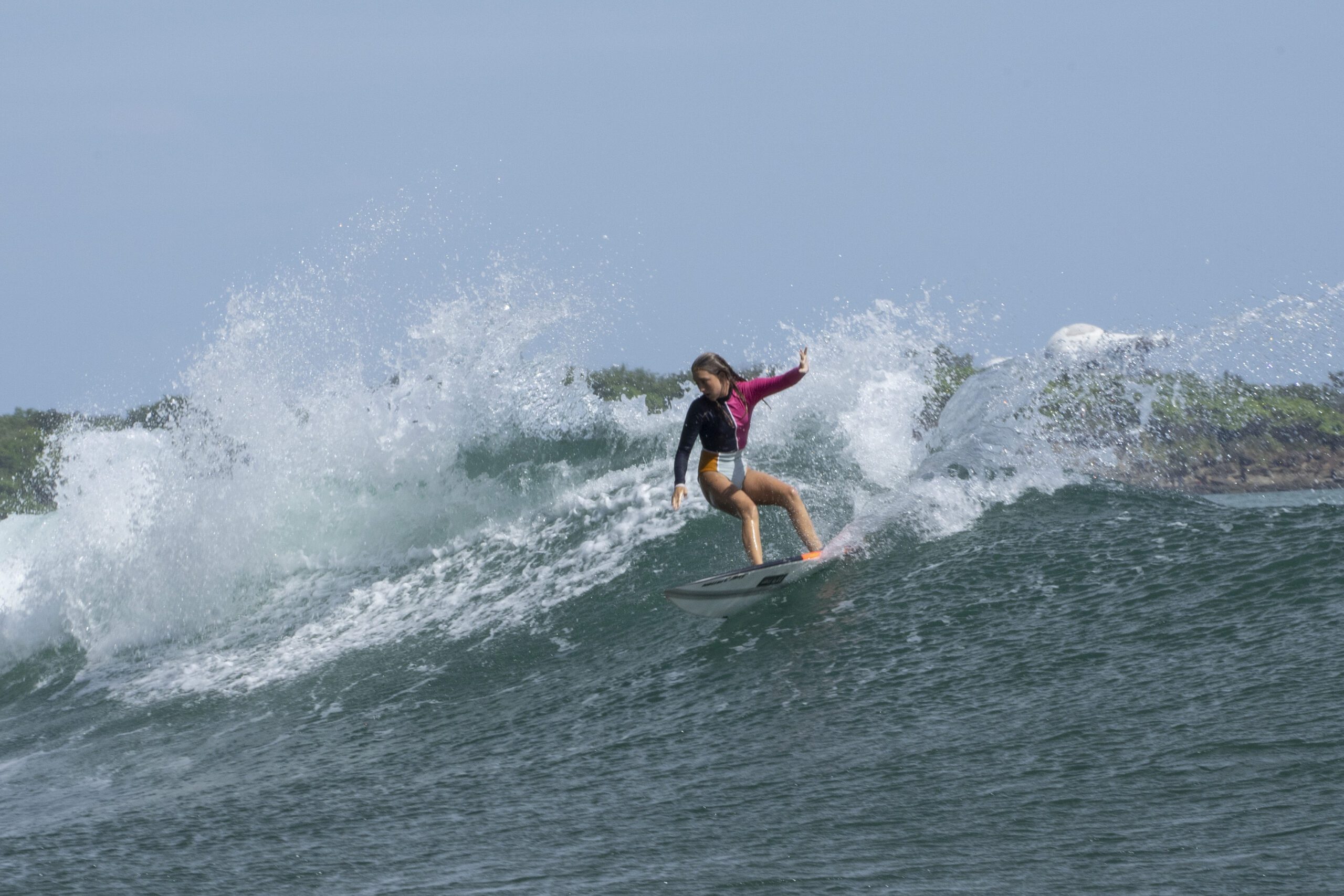 http://Una%20persona%20con%20un%20traje%20de%20neopreno%20rosa%20y%20negro%20monta%20una%20ola%20en%20una%20tabla%20de%20surf,%20con%20agua%20salpicando%20a%20su%20alrededor%20y%20una%20orilla%20verde%20bordeada%20de%20árboles%20al%20fondo,%20bajo%20un%20cielo%20despejado.