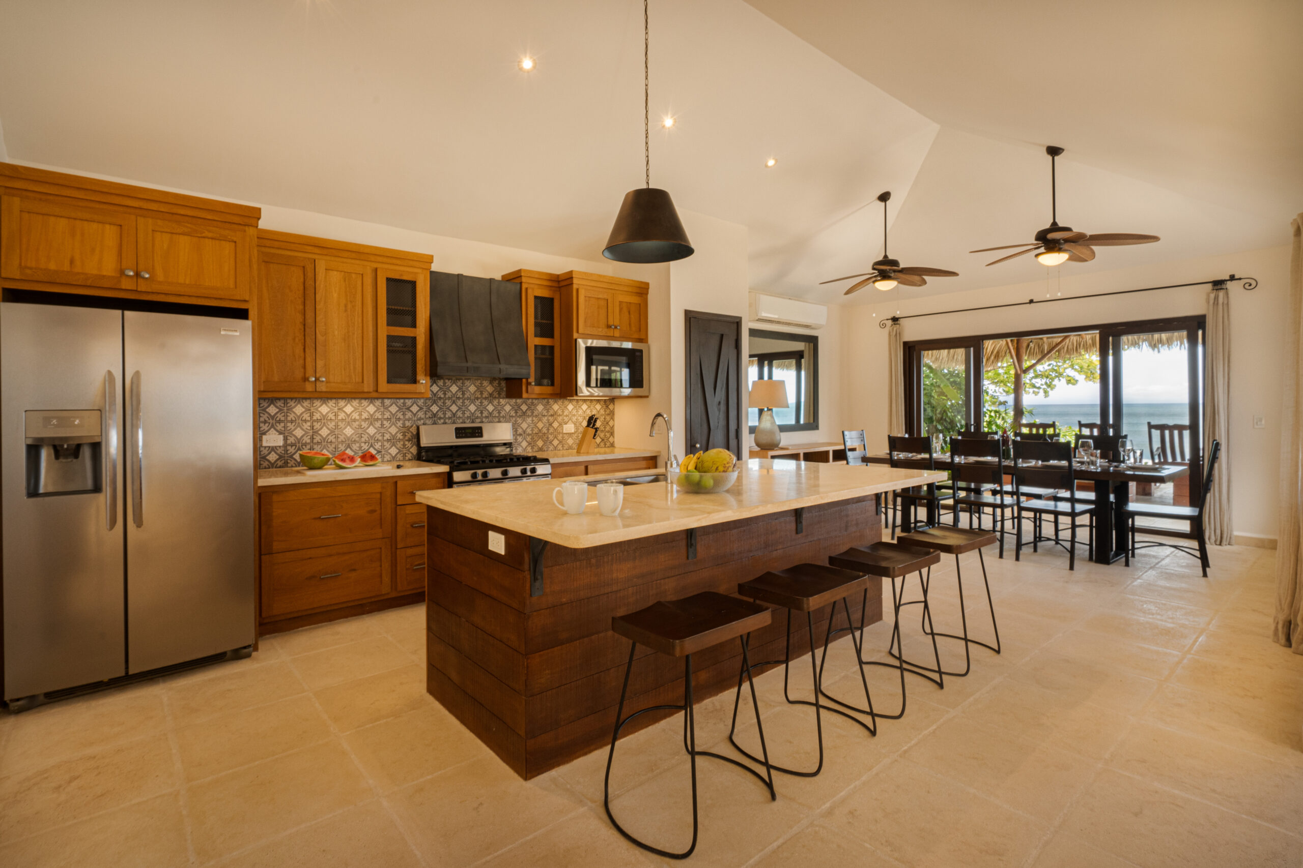http://Modern%20kitchen%20at%20Casa%20Buen%20Día%20featuring%20wooden%20cabinets,%20stainless%20steel%20appliances,%20a%20spacious%20island%20with%20stools,%20pendant%20lights,%20and%20a%20dining%20area%20with%20large%20windows%20overlooking%20a%20tropical%20view.