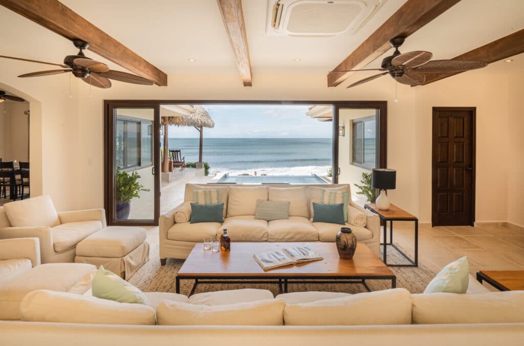 A bright, airy living room at Casa Buen Día features white sofas, wooden beams, ceiling fans, and sliding glass doors that open to a patio, pool, and a scenic ocean view under a blue sky.