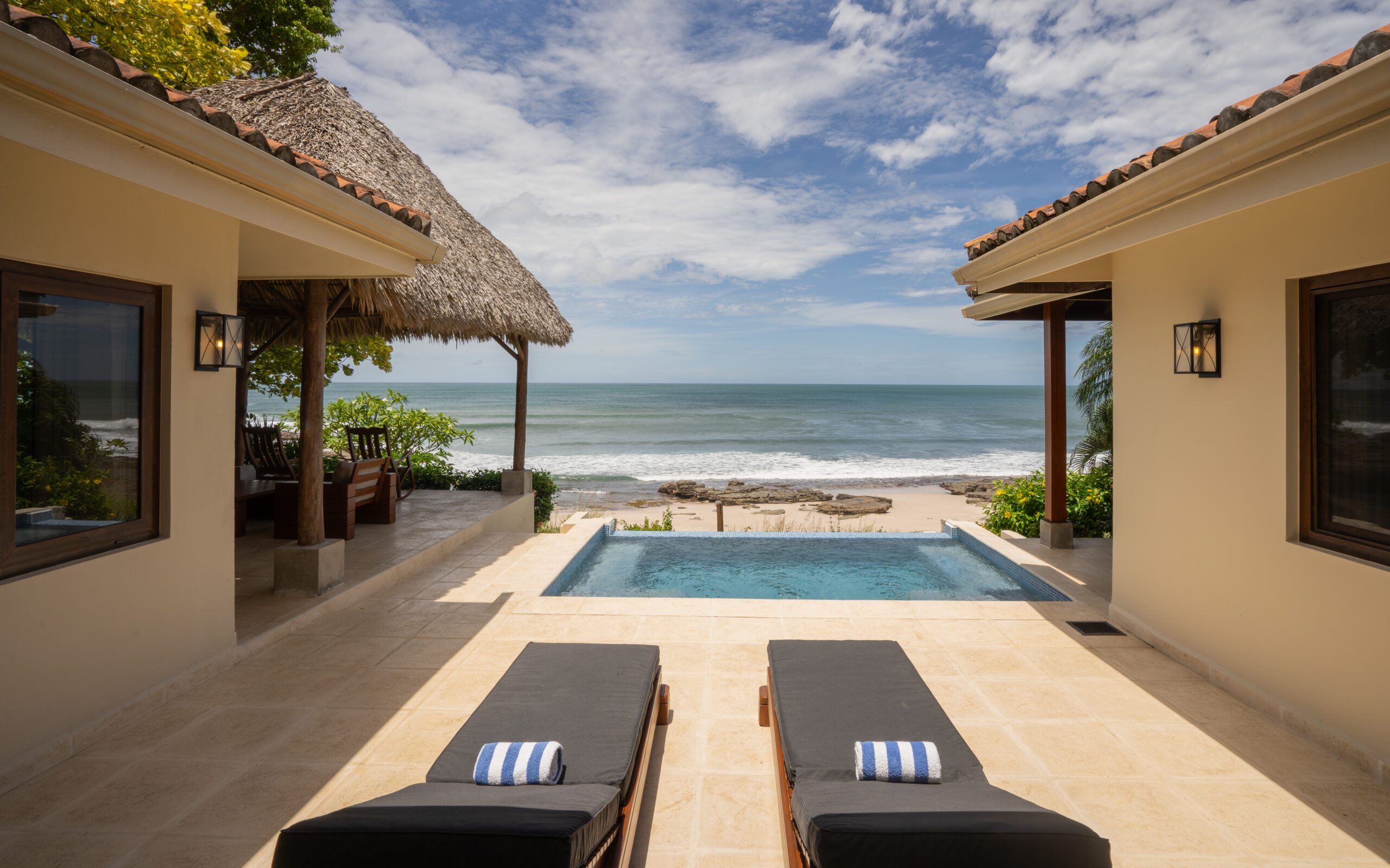 http://Two%20black%20lounge%20chairs%20with%20striped%20towels%20face%20a%20small%20pool%20at%20Casa%20Buen%20Día,%20overlooking%20a%20sandy%20beach%20and%20ocean,%20framed%20by%20two%20beige%20villas%20and%20a%20thatched%20roof%20patio%20under%20a%20partly%20cloudy%20sky.