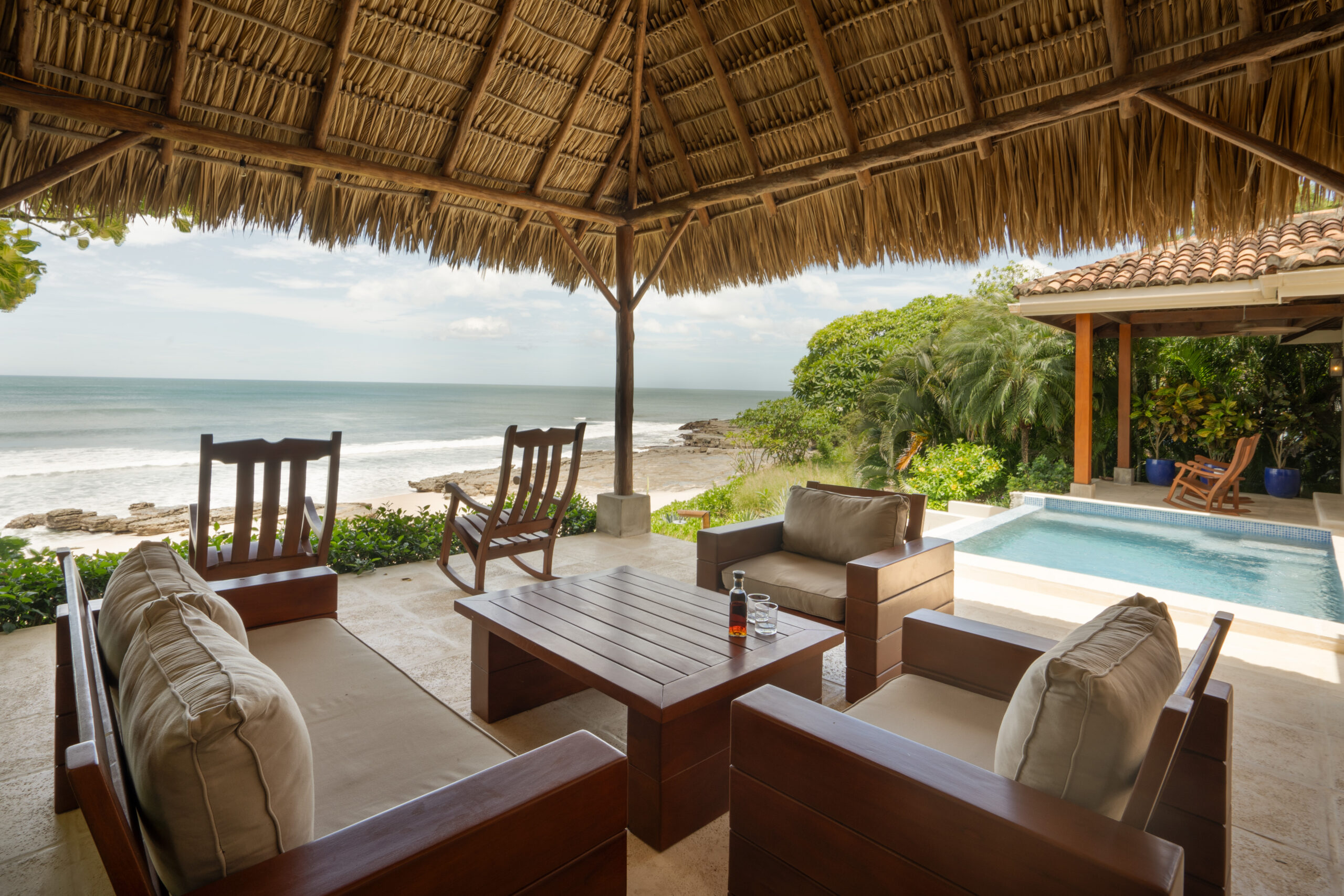 http://A%20shaded%20patio%20at%20Casa%20Buen%20Día%20with%20cushioned%20chairs%20and%20a%20wooden%20table%20overlooks%20a%20sandy%20beach%20and%20ocean.%20A%20small%20pool%20and%20lush%20greenery%20are%20nearby,%20creating%20a%20relaxing,%20tropical%20setting%20under%20a%20thatched%20roof.