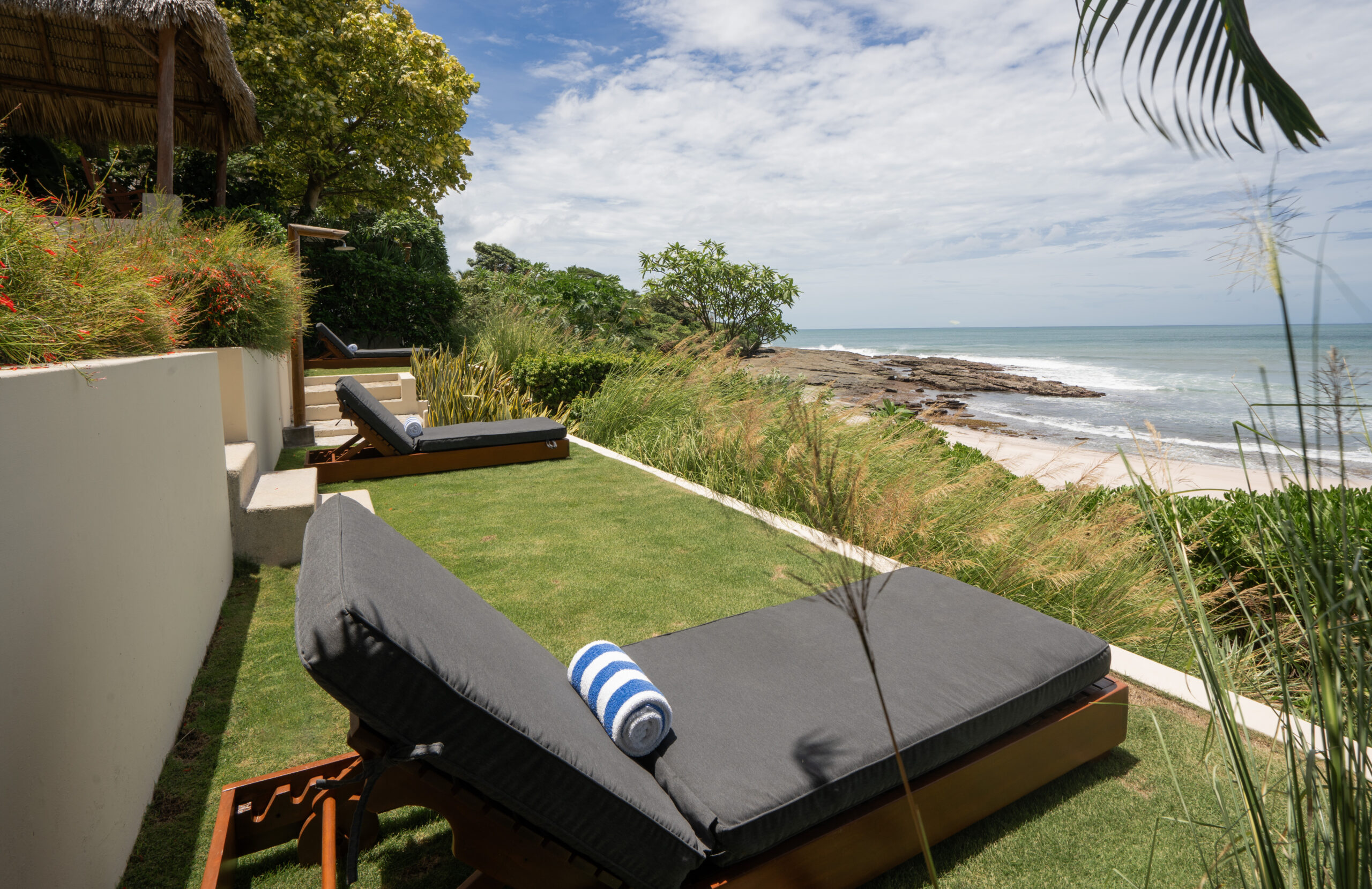 http://Two%20cushioned%20lounge%20chairs%20with%20rolled%20towels%20sit%20on%20a%20grassy%20terrace%20at%20Casa%20Buen%20Día,%20overlooking%20a%20rocky%20shoreline%20and%20the%20ocean,%20surrounded%20by%20lush%20plants%20under%20a%20partly%20cloudy%20sky.