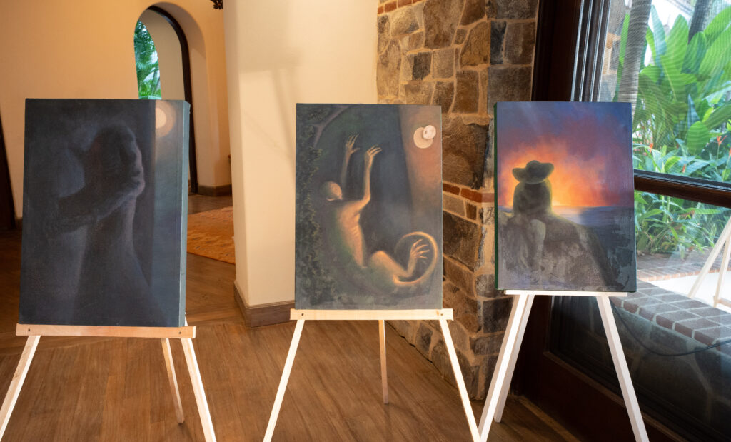 Three paintings on easels indoors: two feature shadowy, ethereal figures with glowing highlights; the third shows a person in a hat sitting on rocks, giving thanks as they gaze at a vivid sunset over the ocean—an earthly paradise.