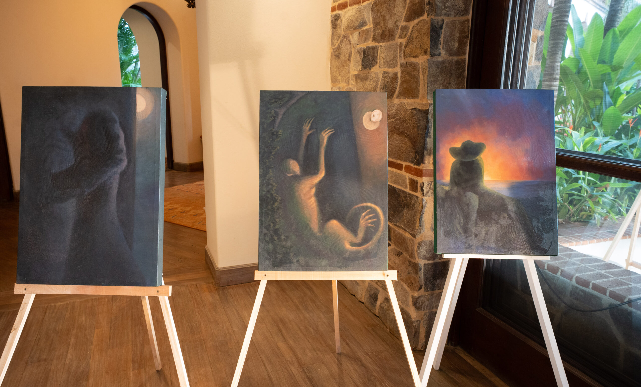 Three paintings on easels indoors: two feature shadowy, ethereal figures with glowing highlights; the third shows a person in a hat sitting on rocks, giving thanks as they gaze at a vivid sunset over the ocean—an earthly paradise.