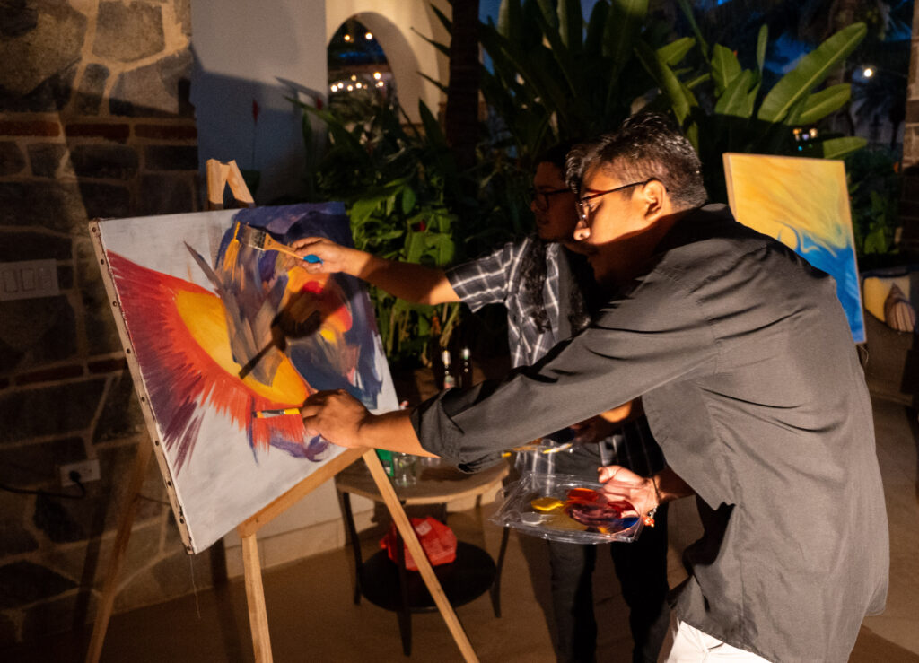 Two men paint together on a canvas at night, using brushes and bright colors. Surrounded by plants and warm lighting in an outdoor paradise, they are focused on their work, giving thanks for the creative moment they share.