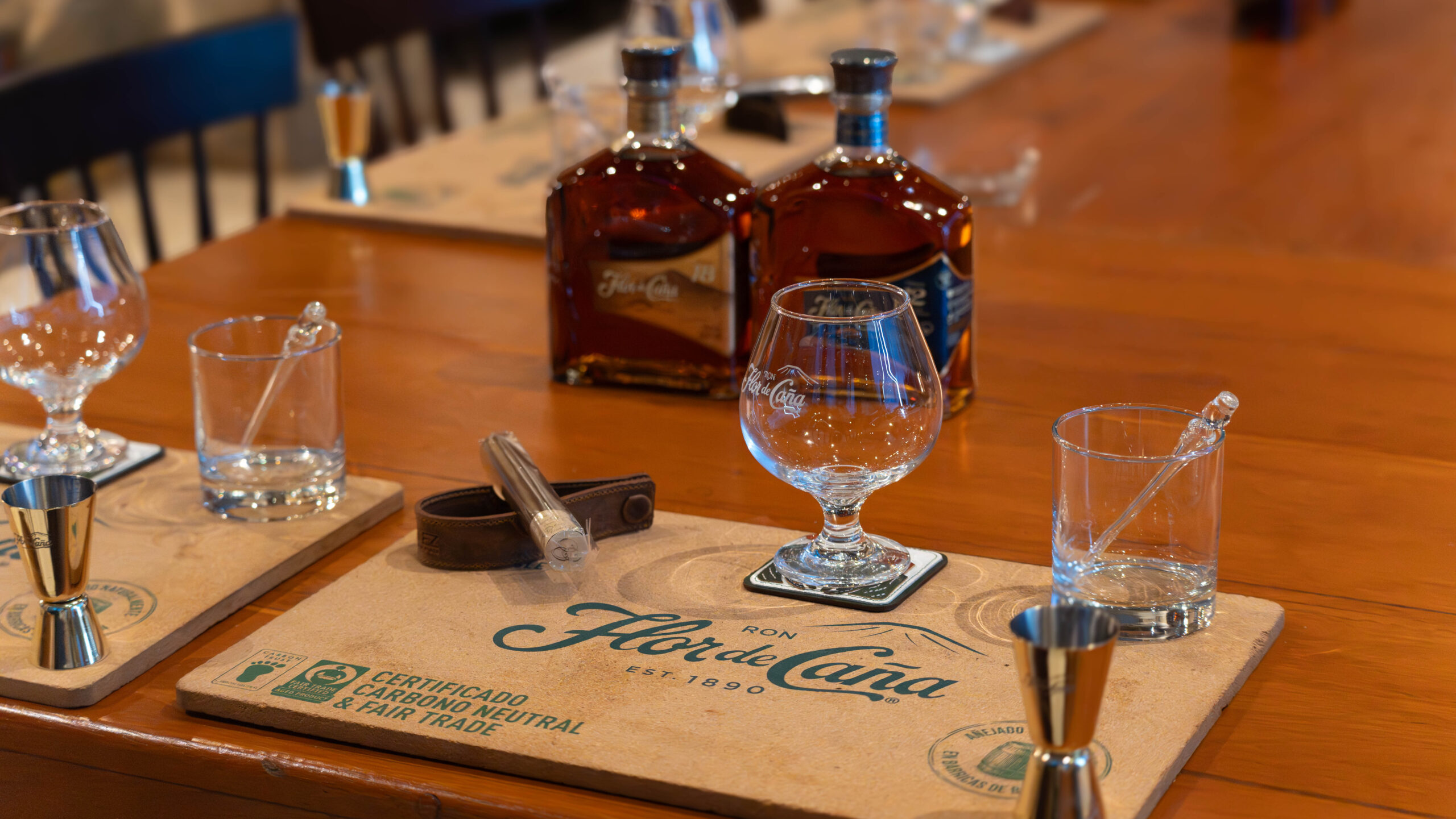 A wooden table set for a rum tasting, featuring Flor de Caña branded placemats, empty glasses, metal jiggers, two bottles of rum, glass stirrers, and a cigar holder with a cigar—true paradise to gather and give thanks.