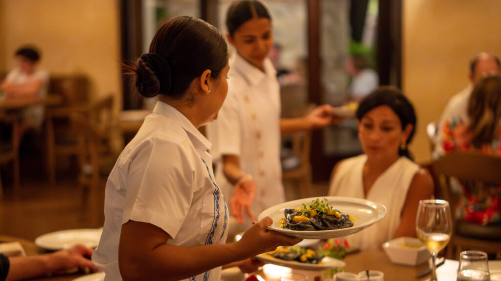 A server in a white uniform hands a plate of food to a seated woman in a restaurant, where the holiday spirit fills the air. Other diners and another server are visible in the warmly lit background.