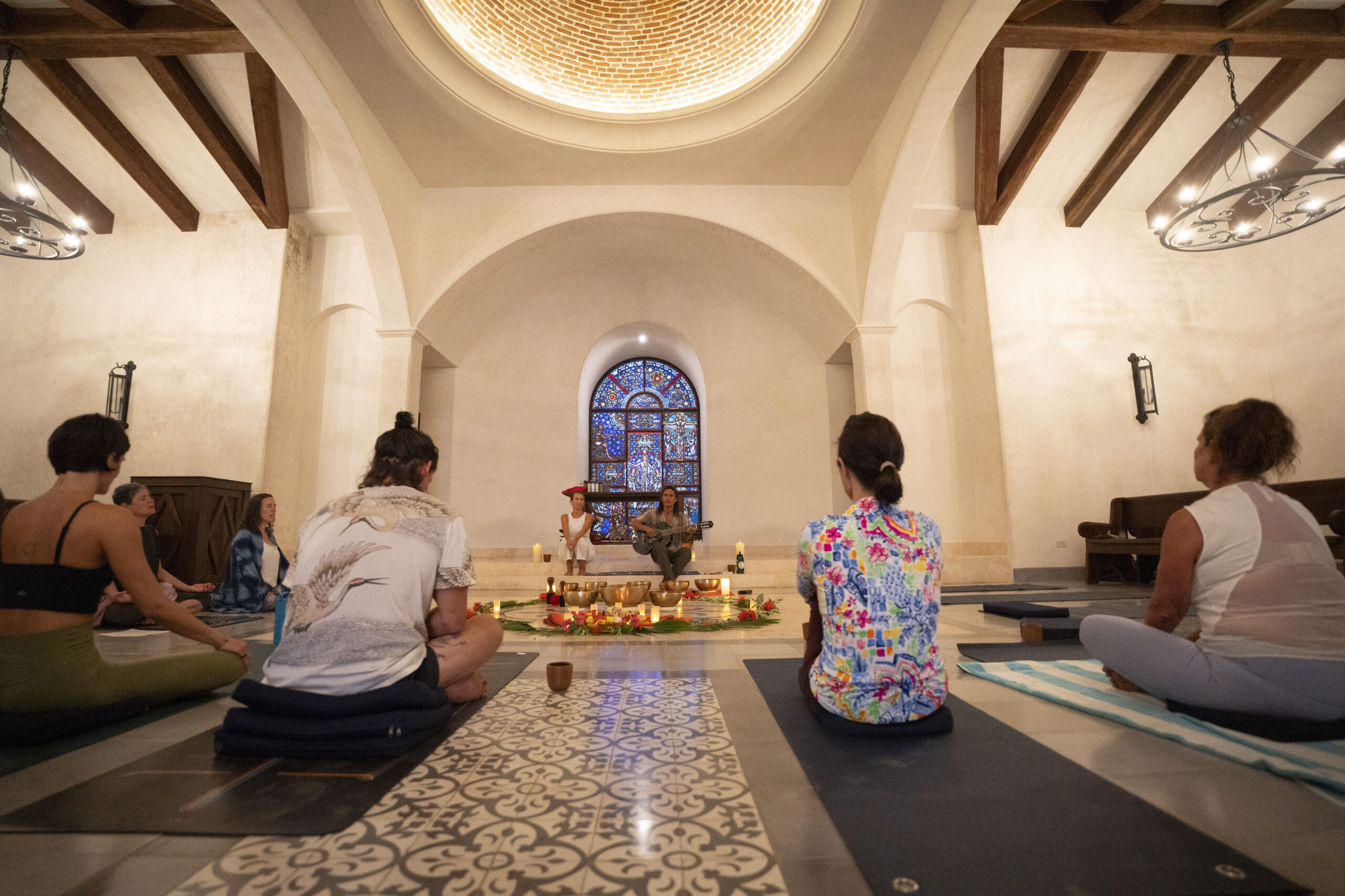 A group of people sit cross-legged on mats in a spacious, warmly-lit room with arched ceilings and stained glass, gathered in a circle around a central altar decorated with candles and flowers, embracing the Holiday Spirit.