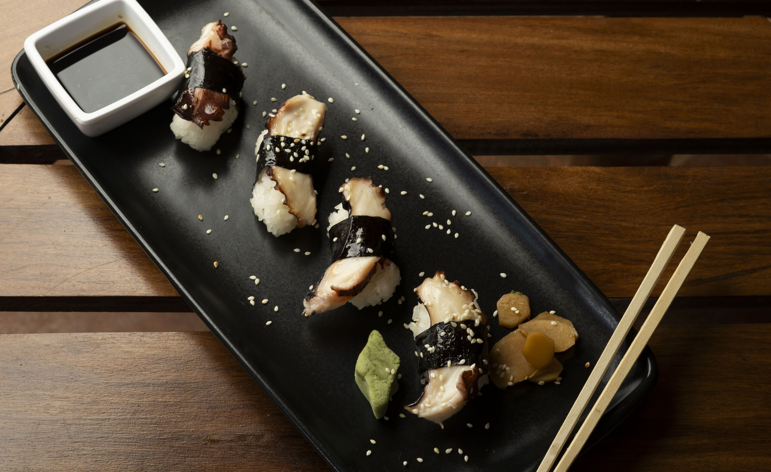 A black rectangular plate with four pieces of octopus nigiri sushi, a small dish of soy sauce, pickled ginger, wasabi, and chopsticks evokes Emerald Coast charm, all set on a wooden table.