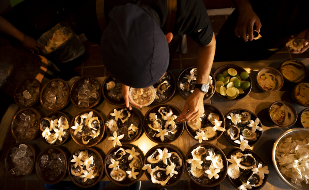 A person arranges white flowers on multiple bowls filled with food or dessert, viewed from above. The table is crowded with dishes, lime halves, and various ingredients, capturing the warm and dim glow of a Holiday Spirit gathering.