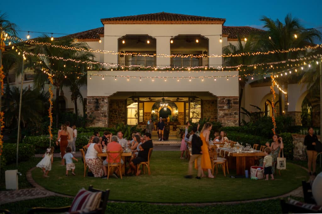 A group of people dine and socialize outdoors on a lawn at dusk, surrounded by string lights, in front of a large, elegant building with palm trees and balconies—capturing the Holiday Spirit along the vibrant Emerald Coast.