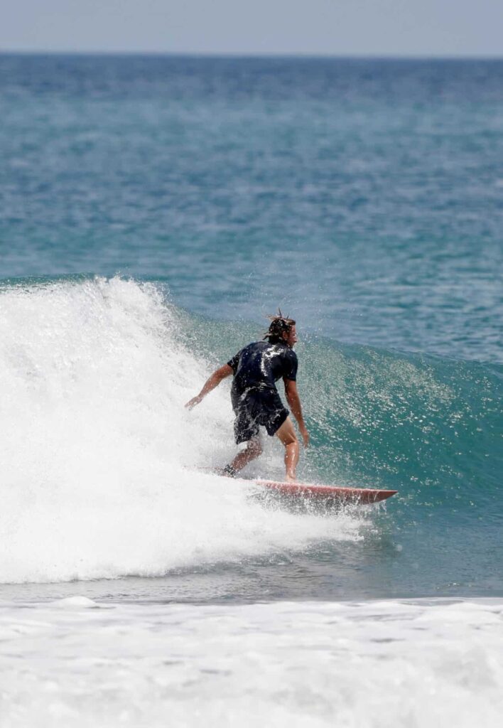 A surfer in a black wetsuit rides a wave on a blue ocean, with white surf trailing behind and a clear sky above.
