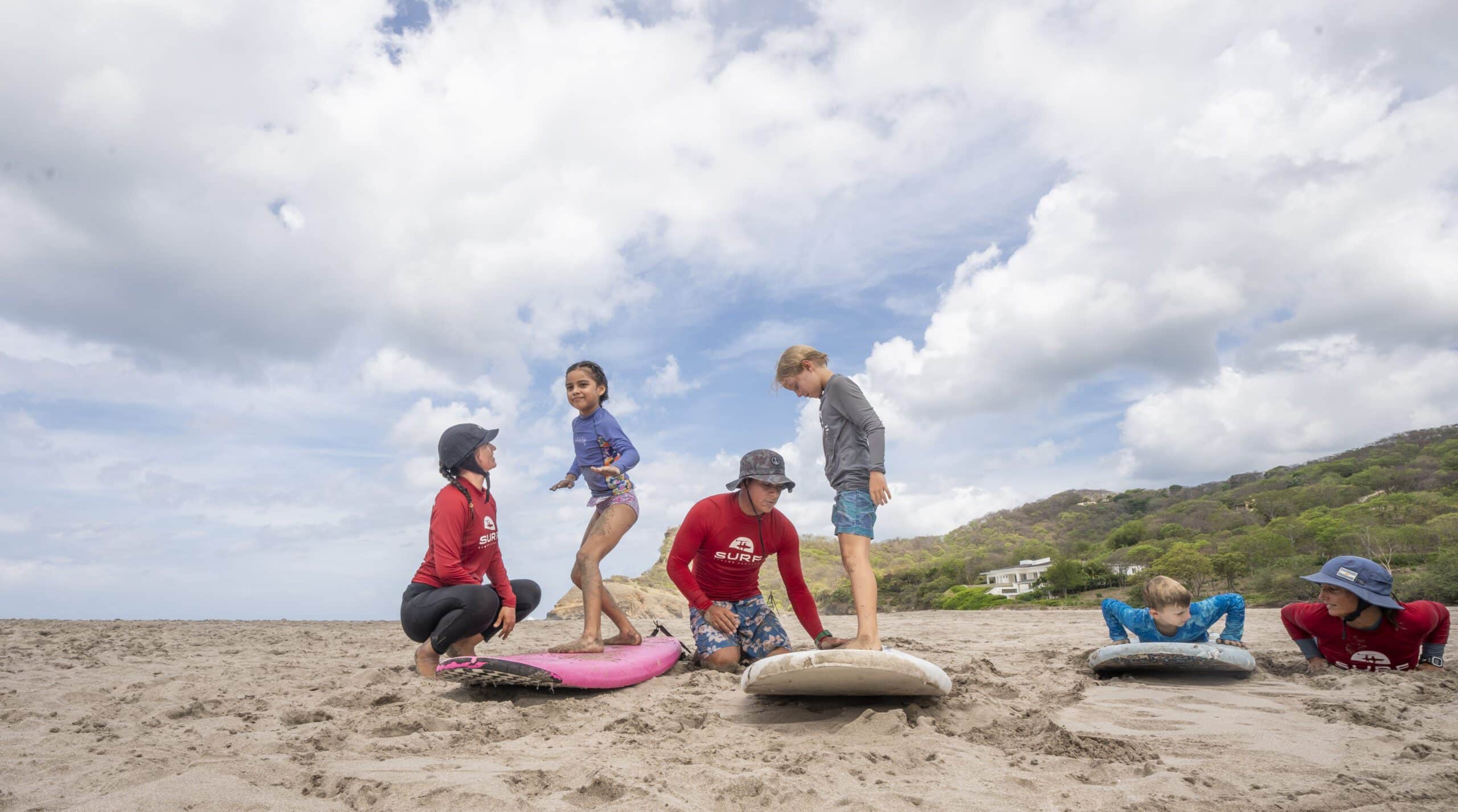 Four children and two instructors practice surfing on the sandy beach under a cloudy sky, ready to catch waves. The instructors kneel beside surfboards while the children stand, kneel, or lay on the boards, mimicking surfing positions and making memories.