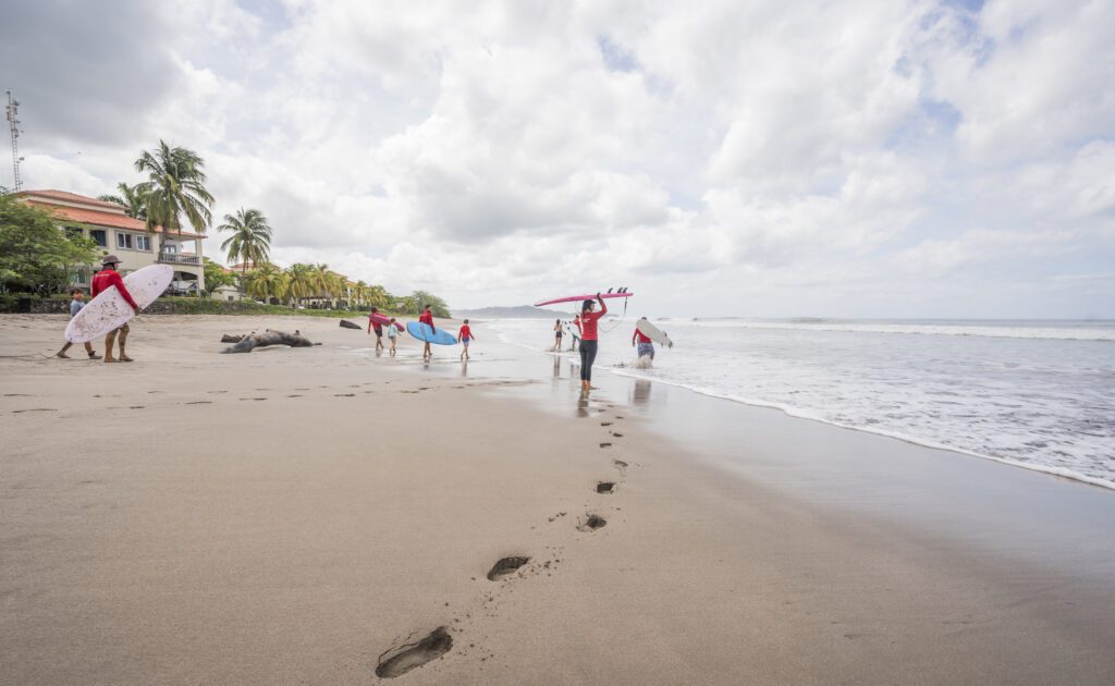 A group of people carrying surfboards walk along a sandy beach toward the ocean, leaving footprints behind as they get ready to catch waves and make memories. Palm trees, buildings, and cloudy skies are visible in the background.