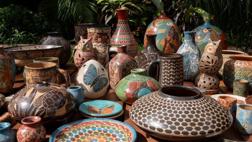 A colorful assortment of handcrafted ceramics by a Chorotegan artisan, including vases, bowls, and plates decorated with animal and nature motifs, displayed on a wooden table outdoors with lush greenery in the background.