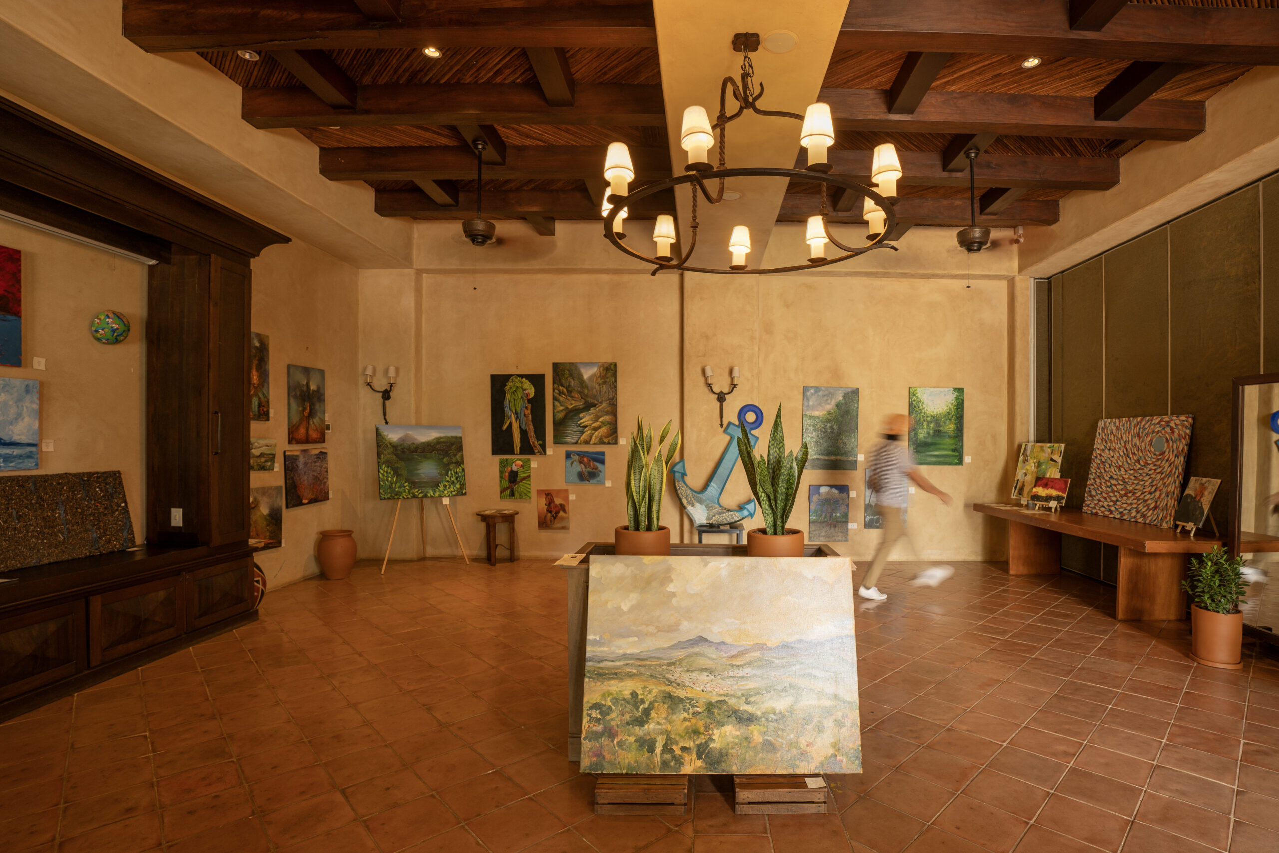 A warmly lit art gallery with rustic decor features landscape and abstract paintings on tan walls and easels. A blurred person walks past the artwork, while potted plants decorate the space during an Exhibición de Arte Nicaragüense.