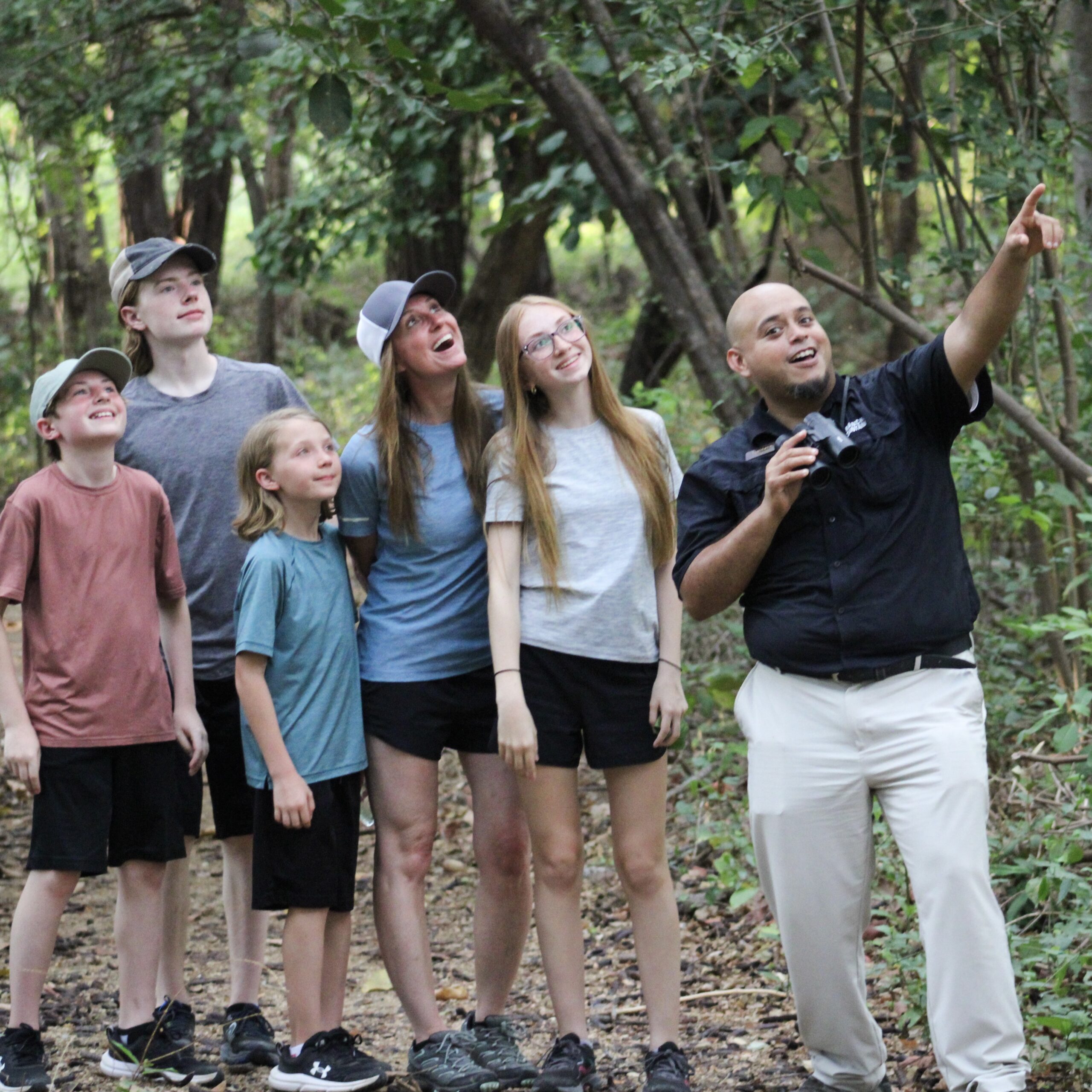 http://A%20group%20of%20five%20people,%20including%20children%20and%20teens,%20stand%20on%20a%20forest%20trail%20while%20a%20man%20in%20a%20black%20shirt%20and%20white%20pants%20points%20and%20talks,%20guiding%20them%20through%20the%20woods.%20Everyone%20looks%20up%20with%20interest.