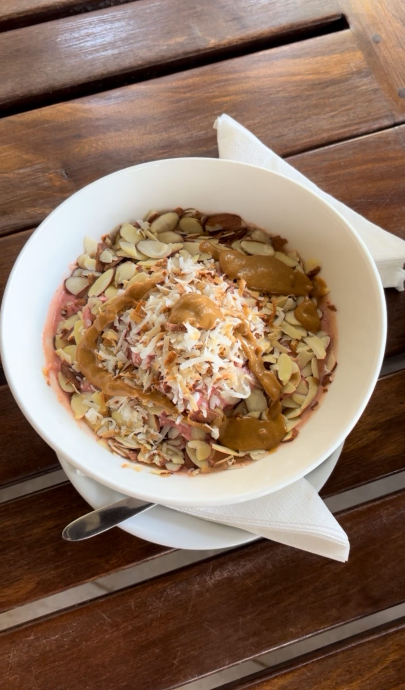 http://A%20white%20bowl%20filled%20with%20a%20smoothie%20topped%20with%20sliced%20almonds,%20shredded%20coconut,%20and%20drizzled%20with%20peanut%20butter,%20placed%20on%20a%20white%20plate%20with%20a%20spoon%20and%20napkin%20on%20a%20wooden%20table.