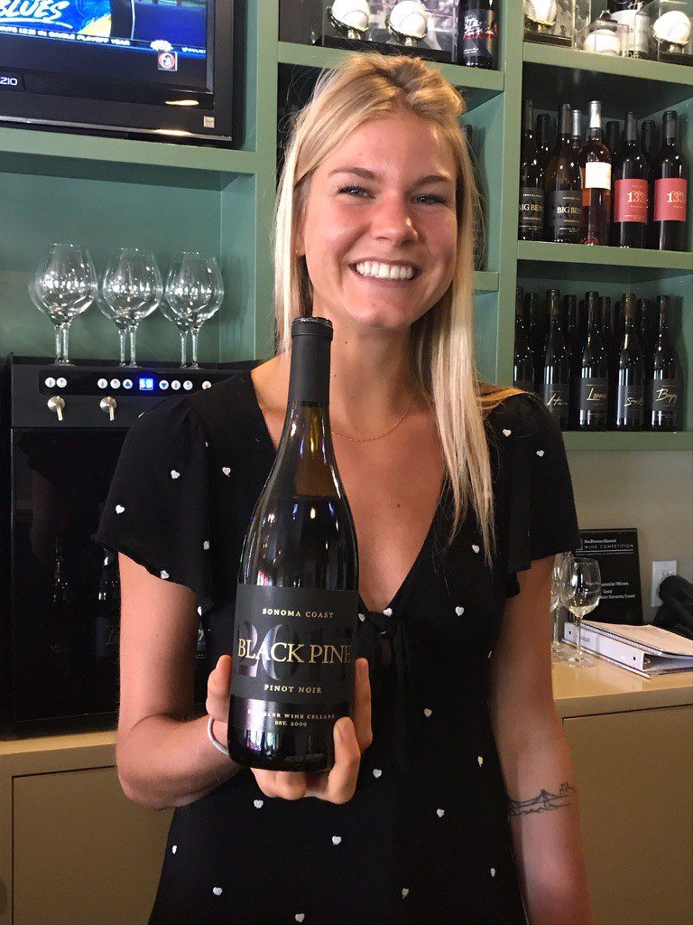 A smiling woman in a black dress with white dots holds a bottle of Black Pine Pinot Noir wine in a wine bar, with shelves of wine bottles and glasses in the background.