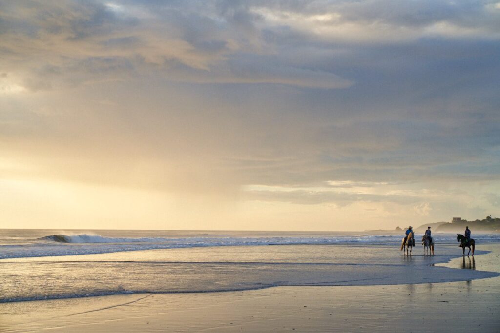 A group of people rides horses along the shoreline at sunset, with gentle waves and a cloudy sky creating a peaceful beach scene.