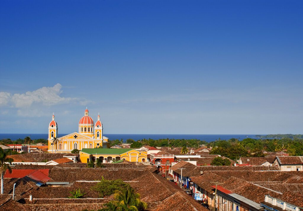 A colorful historic cathedral with red domes rises among red-tiled rooftops and lush greenery, offering a glimpse of a distant blue lake under a clear sky—an iconic gem for culture lovers exploring Nicaragua tourism in Granada.