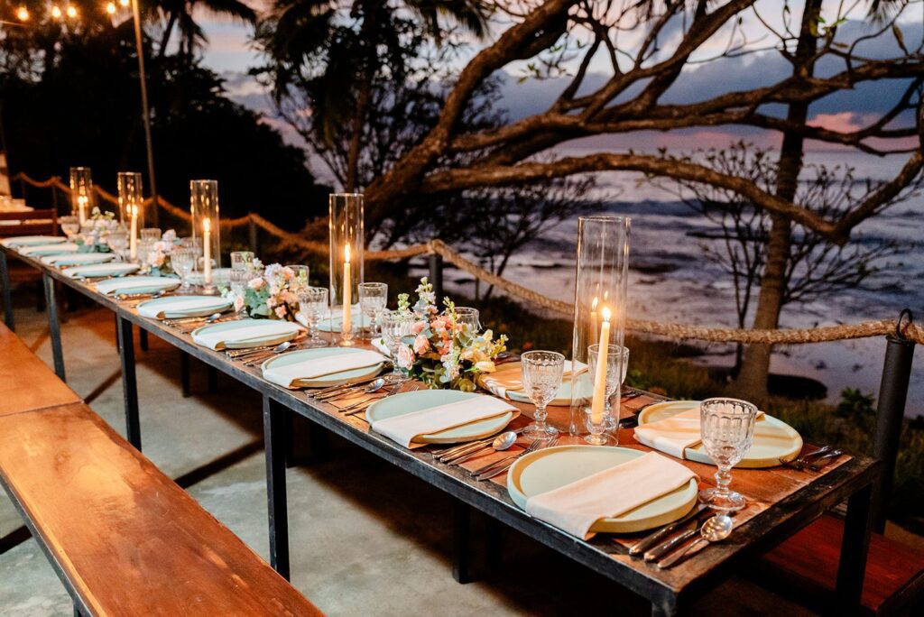 A long outdoor dining table set with plates, glasses, cutlery, candles, and floral centerpieces, overlooking a scenic sunset by the water with trees in the background.