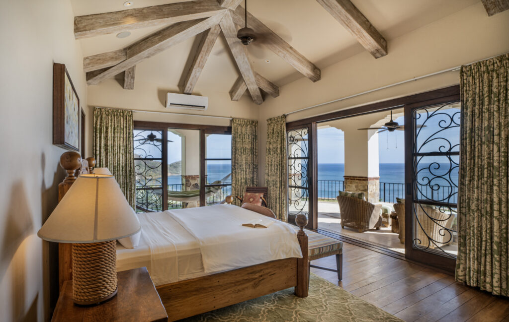 A cozy bedroom in this co-owned luxury home features wooden beams, a large bed, and lamps, opening to a balcony with seating and ocean views. Floor-to-ceiling glass doors let in natural light, framed by patterned curtains at Villa Escondida.