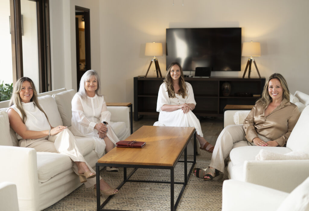 Four women sit comfortably on white couches around a wooden coffee table in a modern living room with neutral decor, large TV, and lamps. Smiling and dressed in light tones, they appear to be brainstorming ideas for corporate event planning.