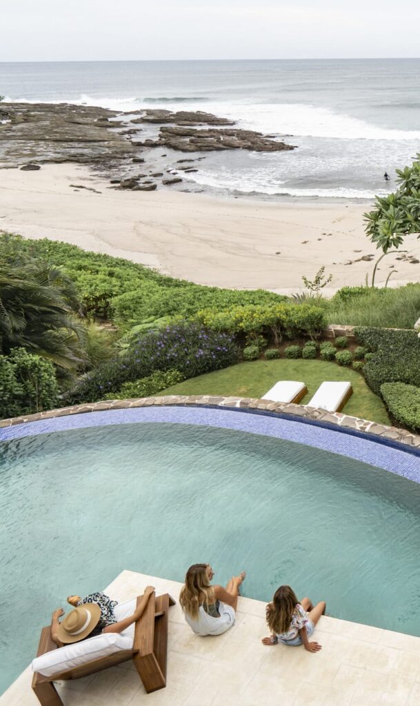 View from above of a tropical beach house: three people relax by a curved pool, while two surfers walk along a path toward the sandy beach and ocean waves.