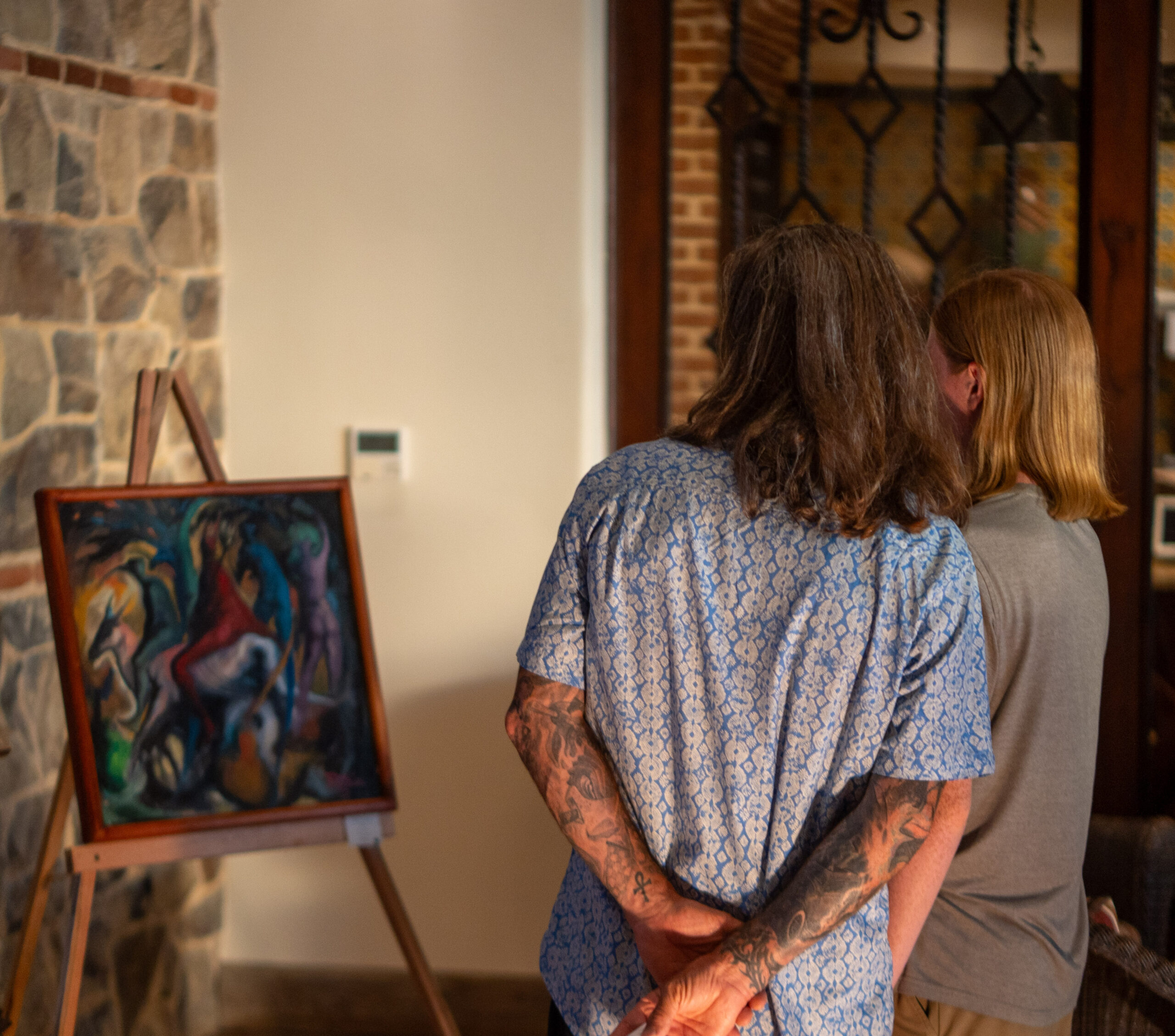 Two people stand closely together, viewed from behind, admiring a colorful abstract painting on an easel inside a room with stone and brick walls at Rancho Santana. One person has visible tattoos and long hair, evoking the creative spirit of Green Season.