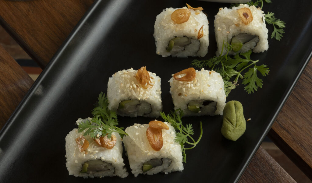 Six pieces of sushi roll topped with fried garlic and sesame seeds, garnished with fresh herbs and wasabi, served on a black rectangular plate—a Green Season favorite at Rancho Santana.