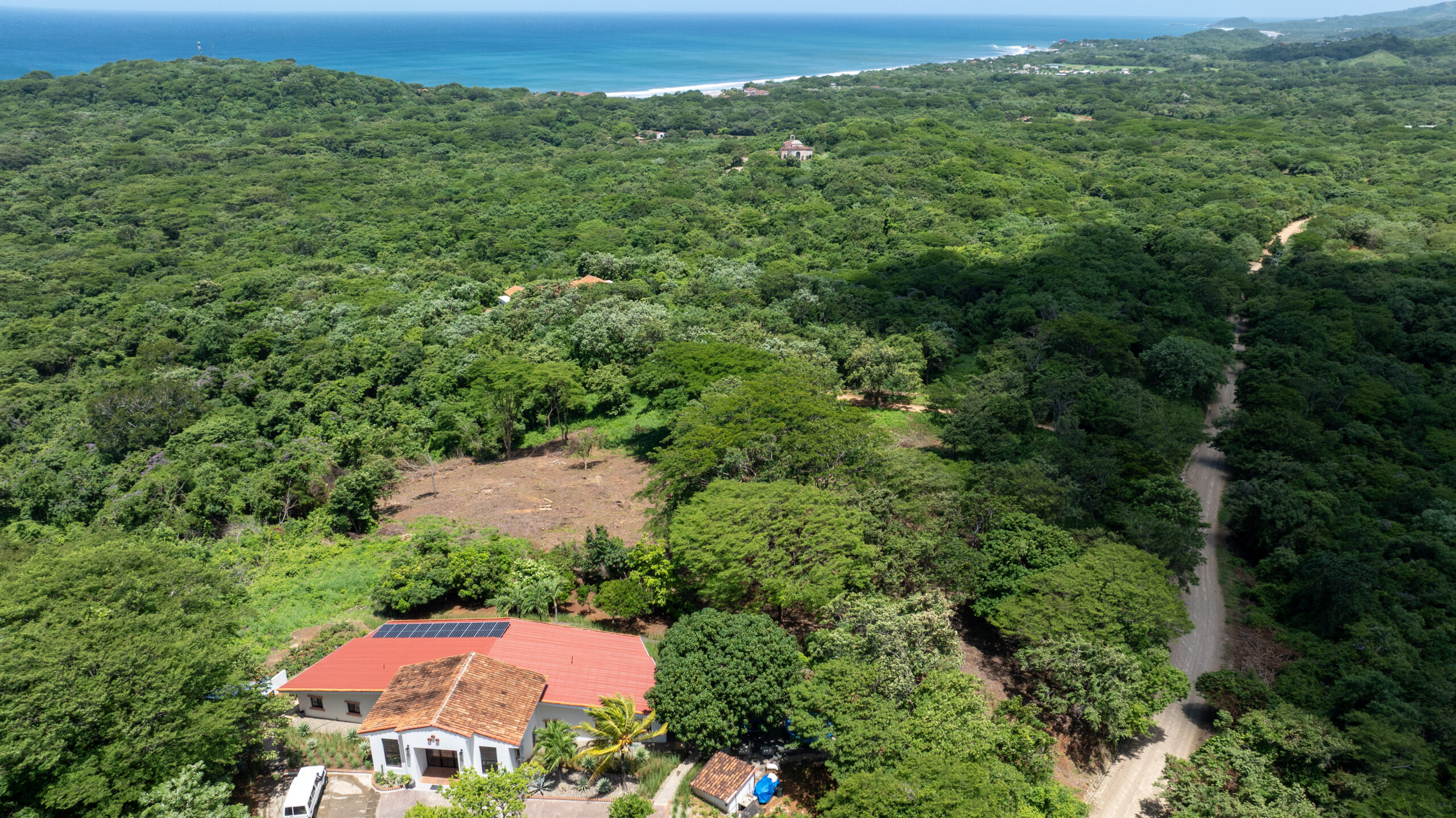 http://Aerial%20view%20of%20a%20house%20with%20a%20red%20roof%20and%20solar%20panels%20surrounded%20by%20dense%20green%20forest,%20dirt%20roads,%20and%20a%20distant%20coastline%20with%20blue%20ocean%20waves%20under%20a%20clear%20sky.