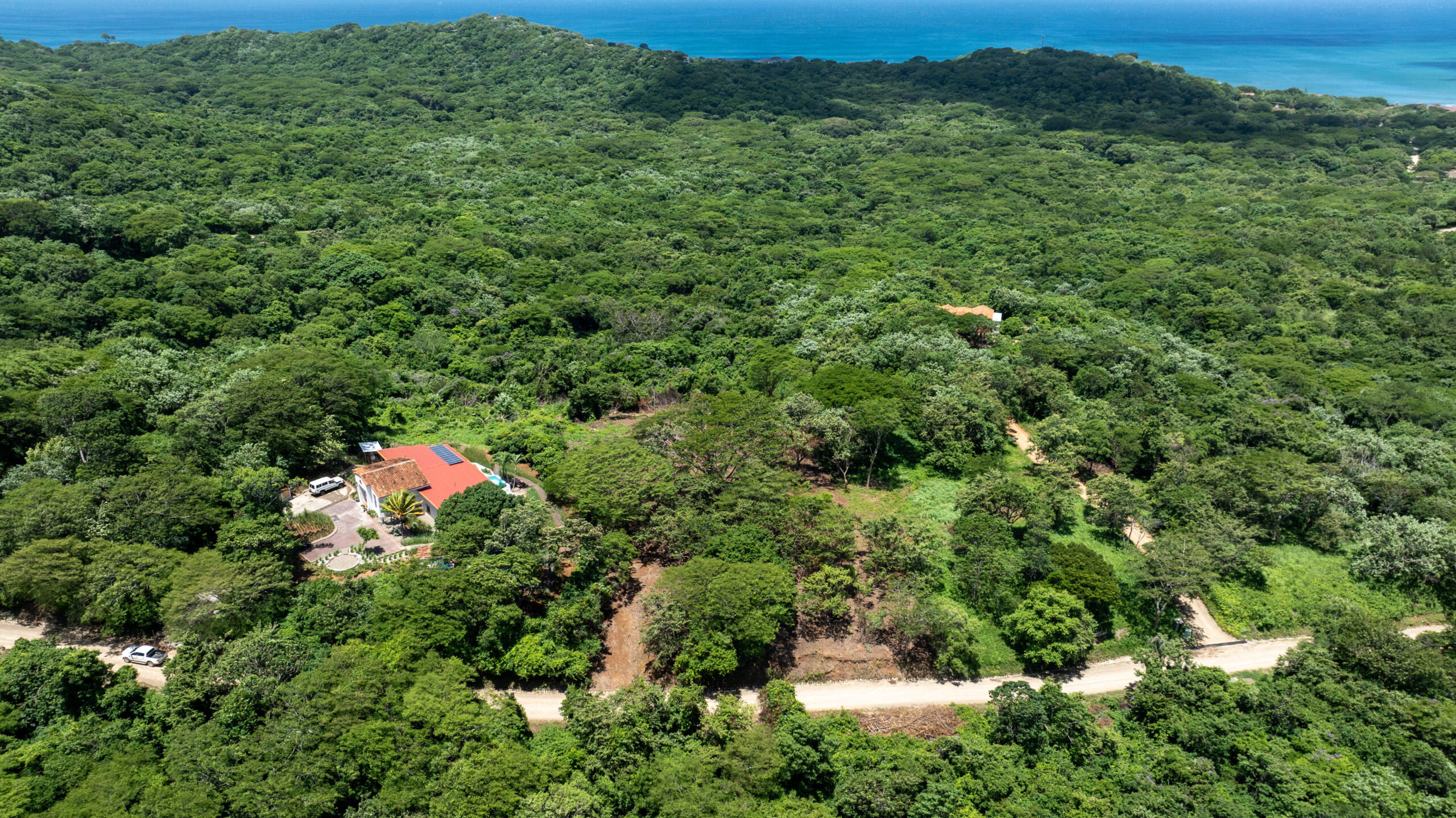 http://Aerial%20view%20of%20a%20lush%20green%20forest%20with%20a%20few%20buildings%20featuring%20red%20roofs,%20a%20dirt%20road%20winding%20through%20the%20trees,%20and%20the%20ocean%20visible%20in%20the%20background%20under%20a%20clear%20sky.
