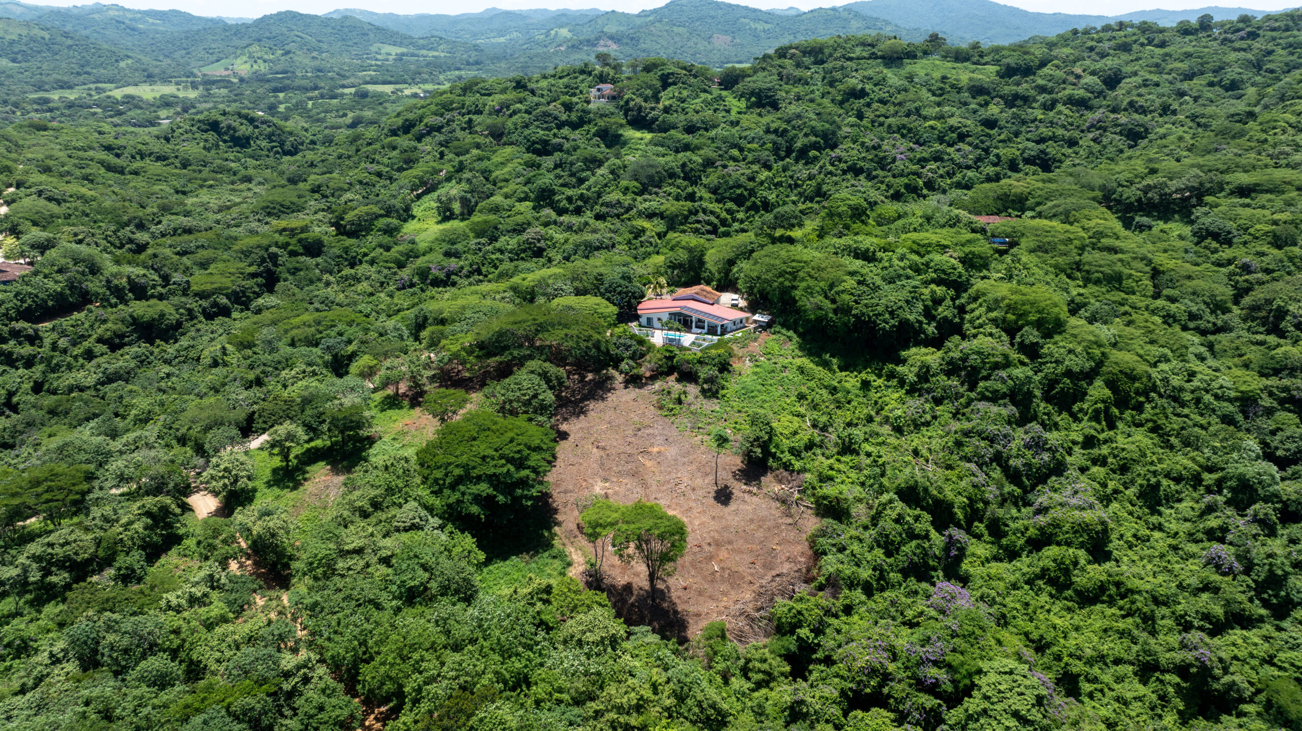 http://A%20large%20house%20with%20a%20red%20roof%20is%20surrounded%20by%20dense%20green%20forest%20on%20a%20hillside,%20with%20a%20cleared%20brown%20patch%20of%20land%20nearby%20and%20mountains%20visible%20in%20the%20distance.